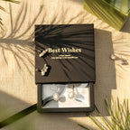 May include: A black gift box with the words "Best Wishes" in gold lettering, containing a pair of silver earrings with a pearlescent center stone and a halo of smaller stones. Two other pairs of earrings are displayed on the box.