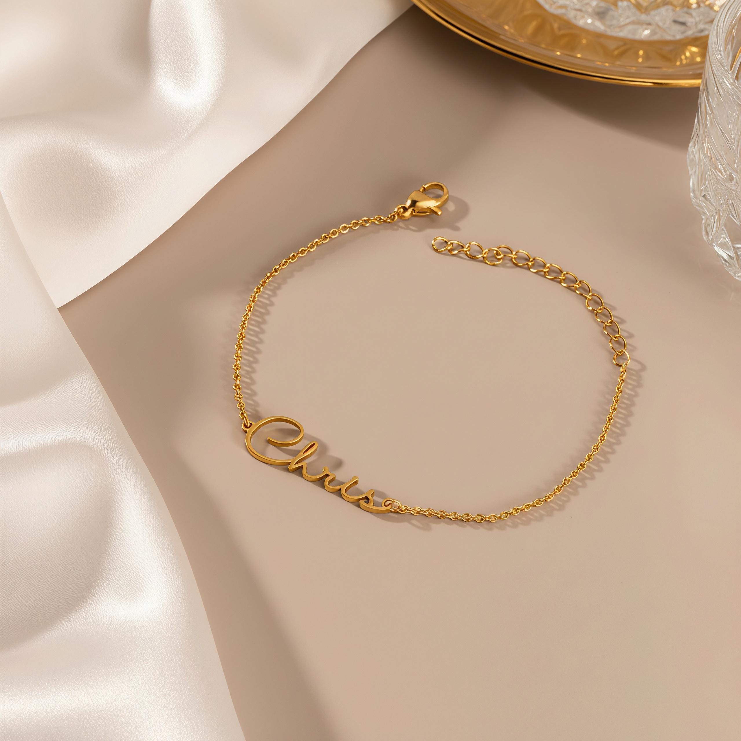 May include: A gold-colored bracelet with the name "Chris" in cursive script. The bracelet is made of a delicate chain and has an adjustable length. The clasp is visible at the end of the chain. The bracelet is displayed on a neutral-toned surface.