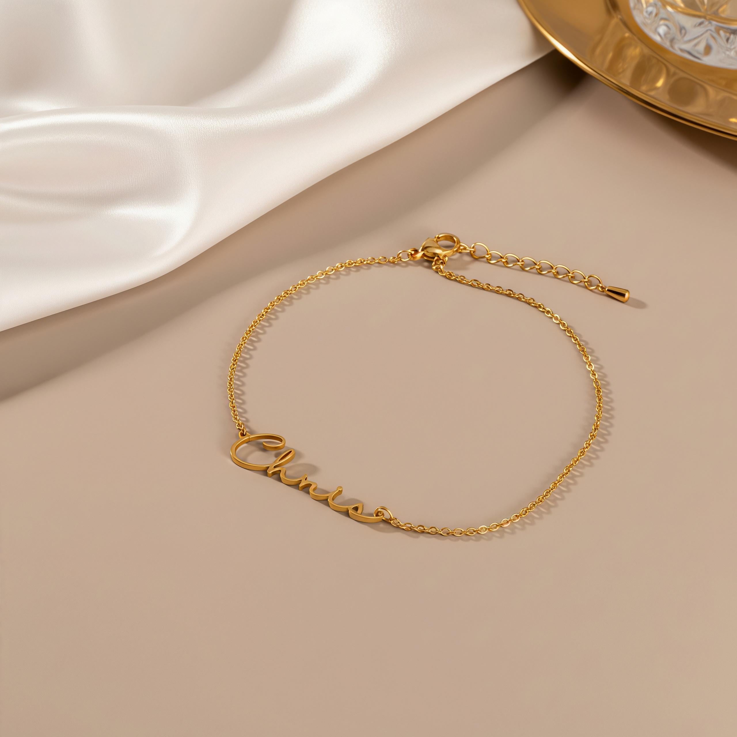 May include: A delicate gold-colored bracelet with the name "Chloe" in cursive script. The bracelet features a fine chain and an adjustable clasp. The jewelry is displayed on a neutral-toned surface, with a soft, white fabric in the background.