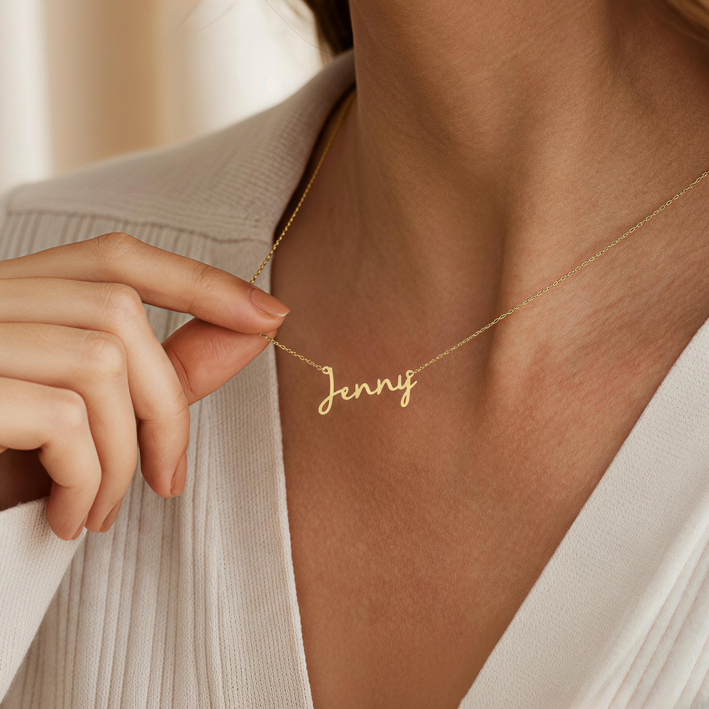 May include: A delicate gold-colored necklace with the name "Jenny" in a cursive script. The necklace is displayed against a neutral background, highlighting the jewelry's elegant design. The chain is thin and the pendant is in a handwritten style.