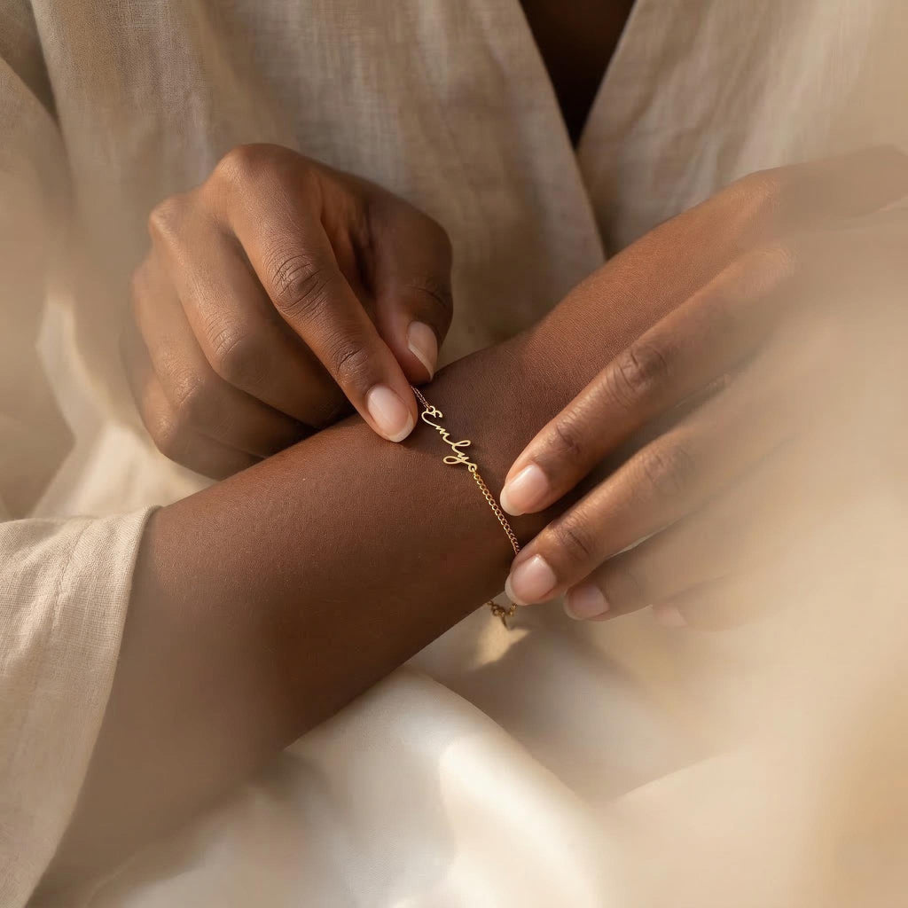 May include: A delicate gold bracelet with the name "Emly" in cursive script. The bracelet is on a slender chain and is being adjusted on a wrist. The background is a soft, neutral color.