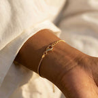 May include: A delicate gold-colored bracelet with a lobster clasp closure. The bracelet is a fine chain design, worn on a wrist. The background is a soft, neutral color.
