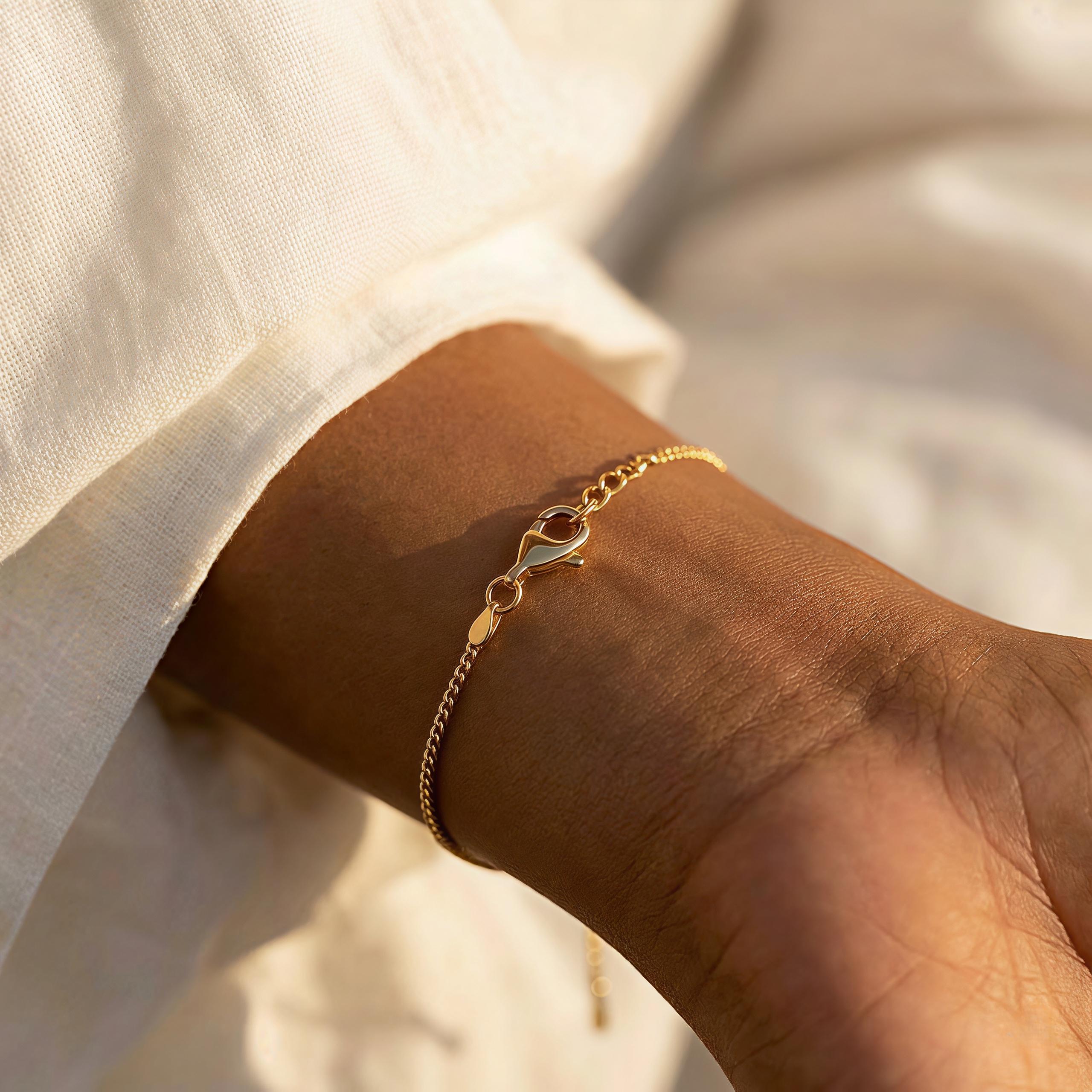 May include: A delicate gold-colored bracelet with a lobster clasp closure. The bracelet is a fine chain design, worn on a wrist. The background is a soft, neutral color.