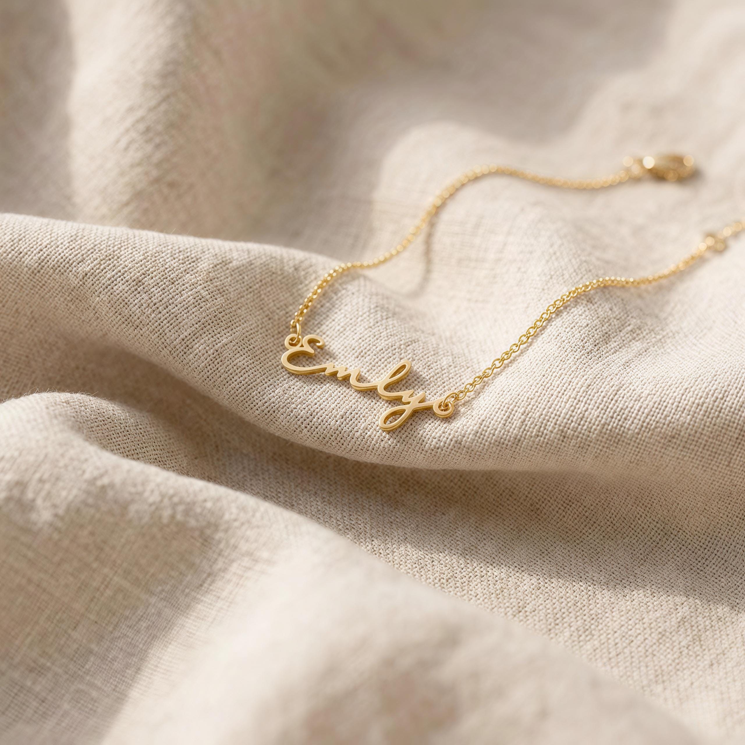 May include: A delicate gold-colored necklace with the name "Emily" in cursive script. The necklace is displayed on a beige, textured fabric, highlighting the jewelry's elegant design. The chain is thin and the clasp is visible.
