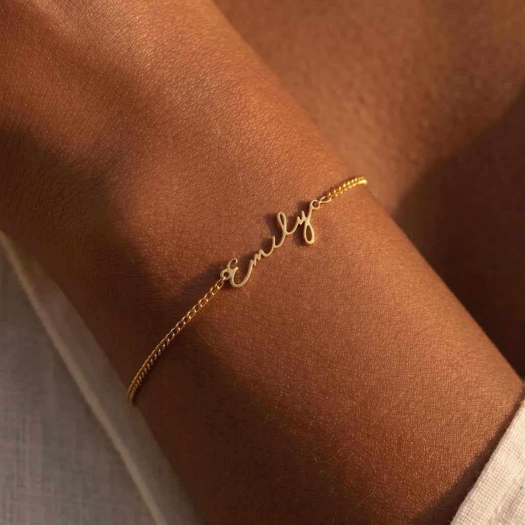 May include: A delicate gold bracelet with the name "Emily" in cursive script. The bracelet is a fine chain, and the name is centered. The image is a close-up, showing the bracelet on a person's arm.