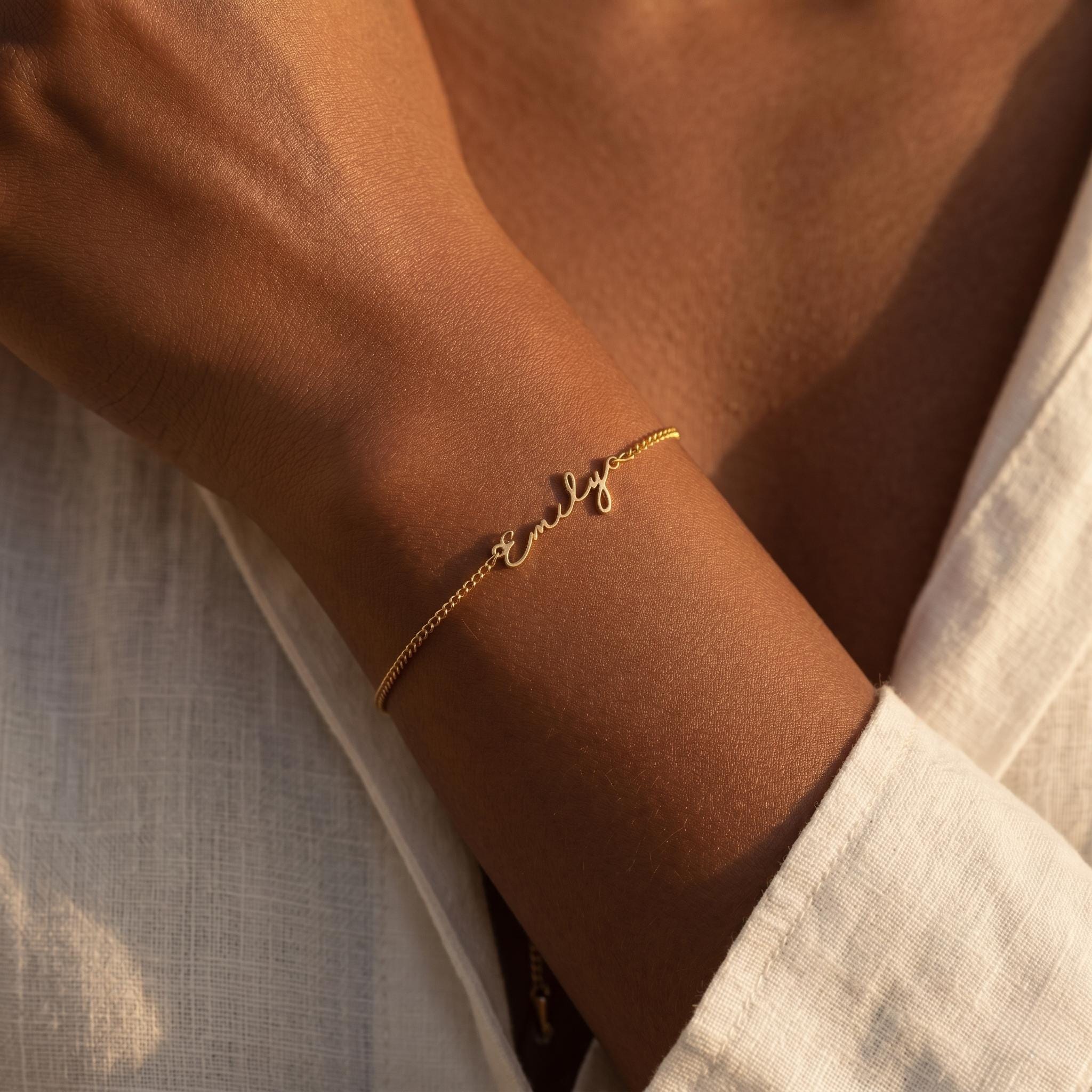 May include: A delicate gold bracelet with the name "Emily" in cursive script. The bracelet is on a slender chain and is worn on a wrist. The background is a white shirt.